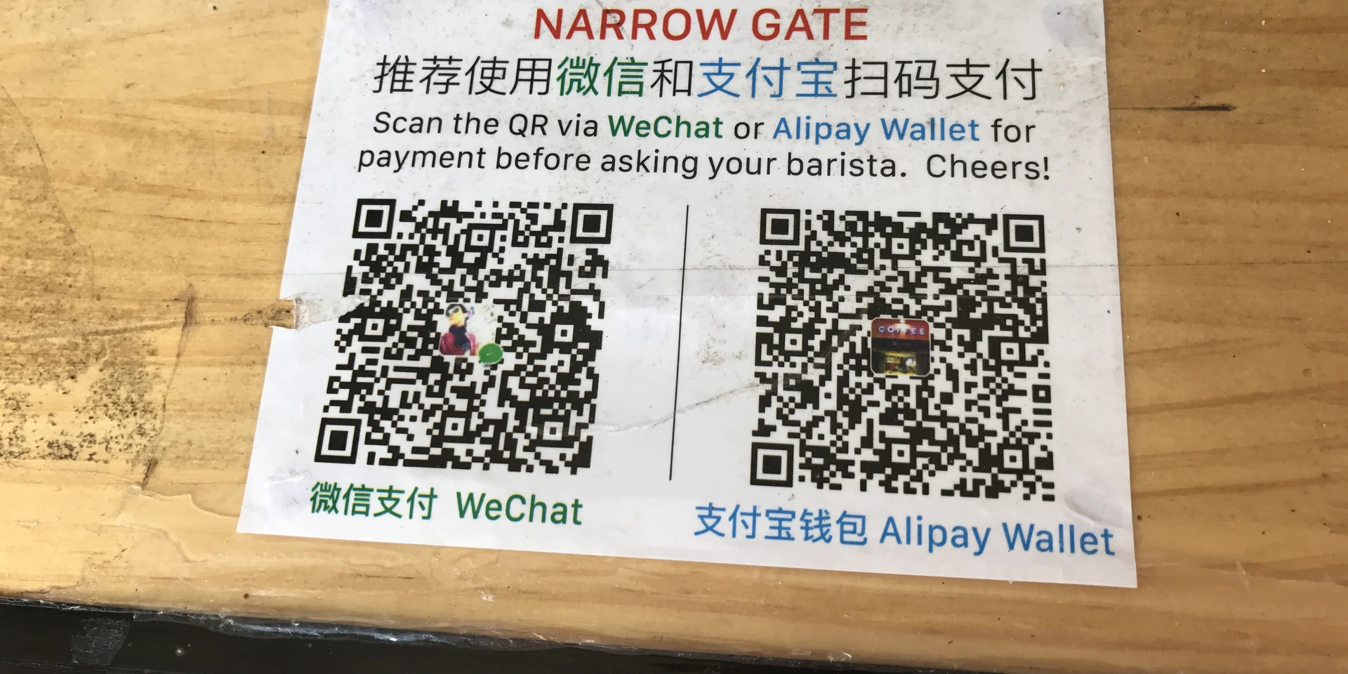 A printed sign on a wooden café counter in China with two payment QR codes, one labelled WeChat and one labelled Alipay Wallet, with the line Scan the QR via WeChat or Alipay Wallet for payment before asking your barista