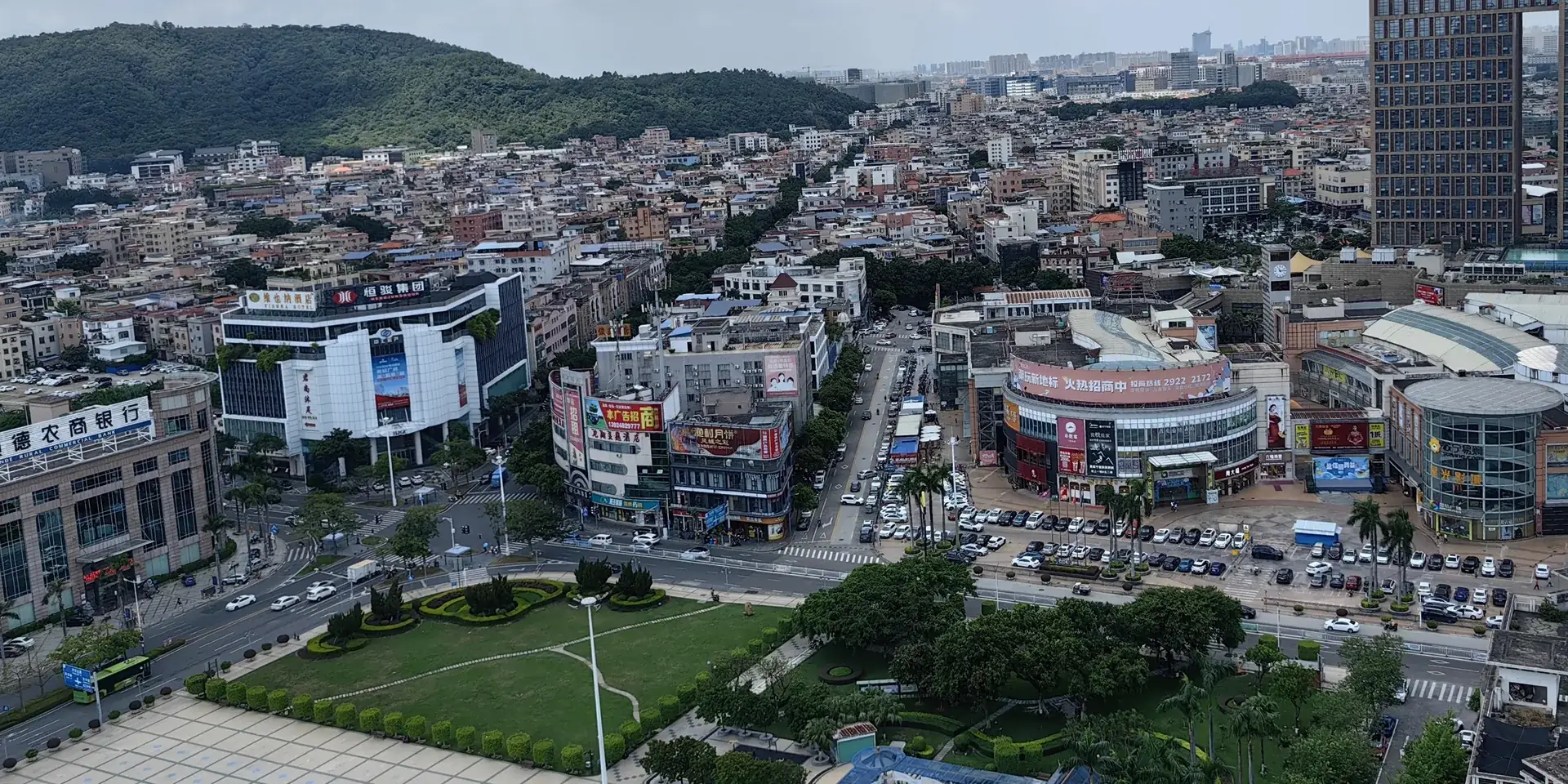 View north over Longjiang town in Shunde, Foshan, from a high window: a central plaza and lawn in front, shopping buildings and dense low-rise blocks behind, and a green hill on the left under a cloudy sky