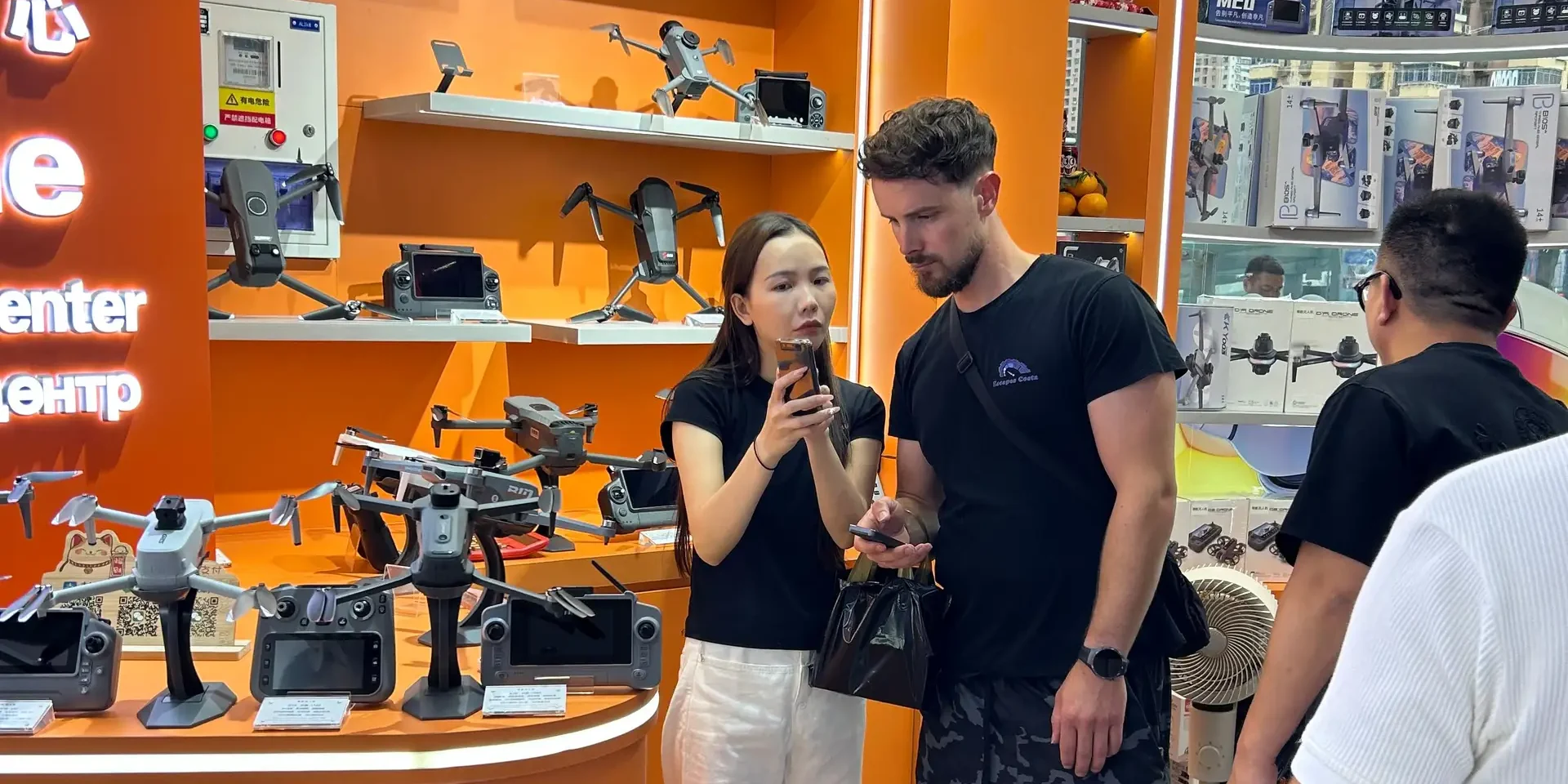 Bilingual interpreter showing a Western buyer product details on her phone inside a Huaqiangbei drone wholesale shop, with drones on orange shelves